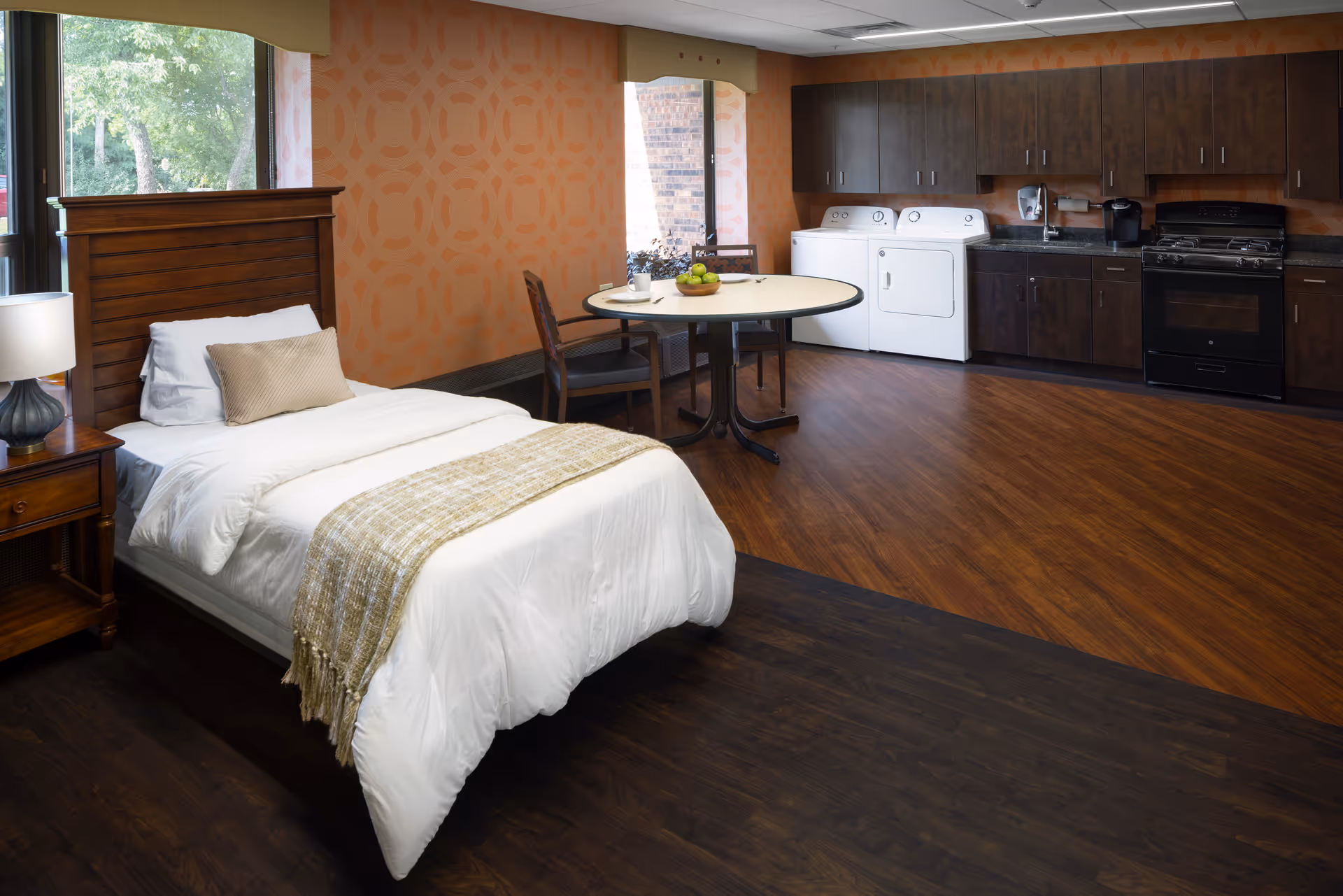 A senior living facility room featuring a single bed with white bedding and a beige throw blanket, a wooden nightstand with a lamp, a round dining table with two chairs, and a kitchenette area with dark wood cabinets, a stove, a sink, a coffee maker, and a washer and dryer. The room has large windows with views of greenery outside and patterned orange wallpaper.