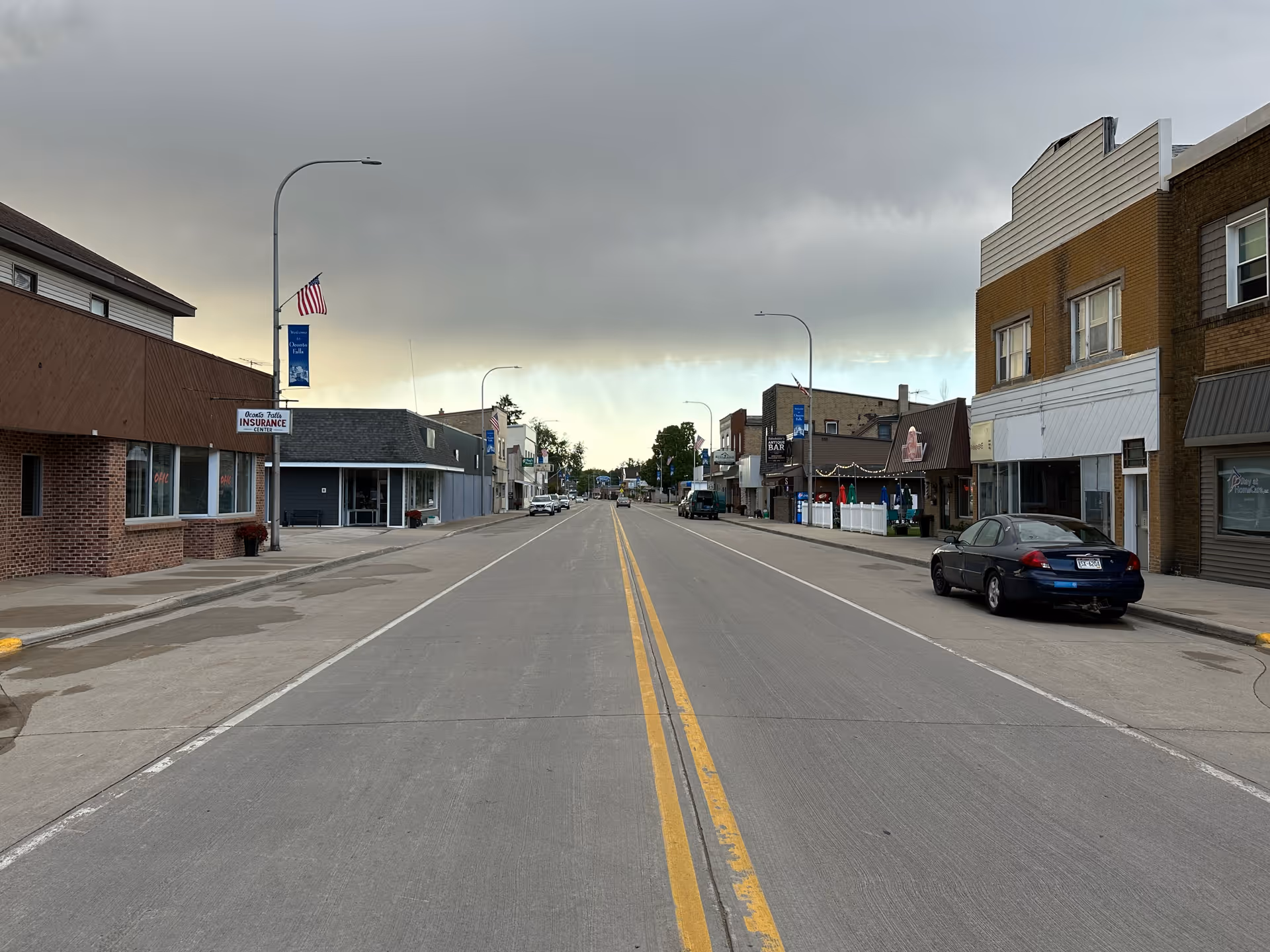 A quiet downtown street with storefronts on both sides beneath a cloudy sky.