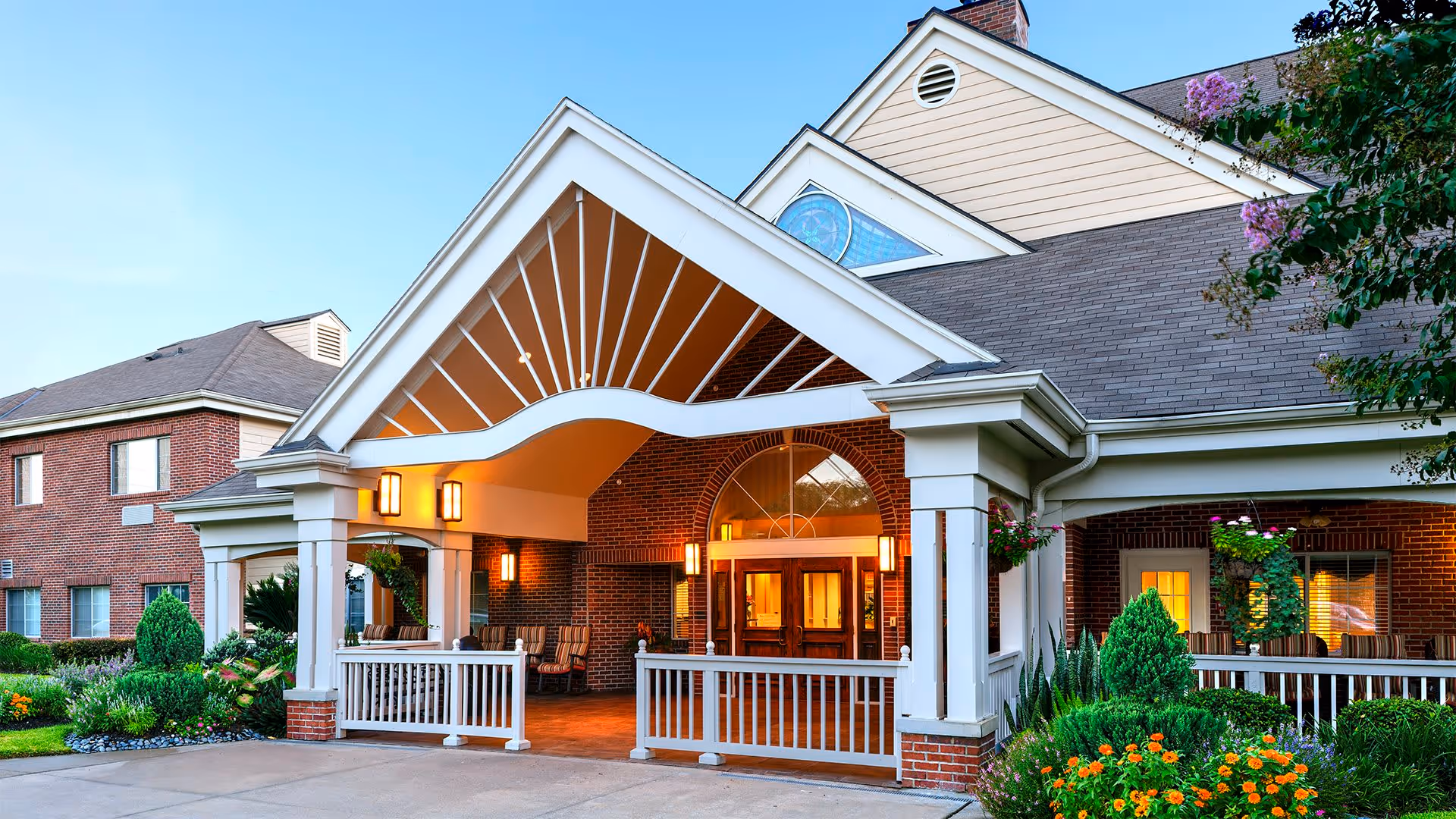 Covered entrance and porte-cochère of a brick senior living building with white columns, lit entryway, and landscaped flowerbeds.