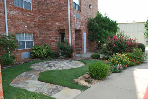 Brick building entrance with a stone walkway, green lawn and landscaped flowerbeds.