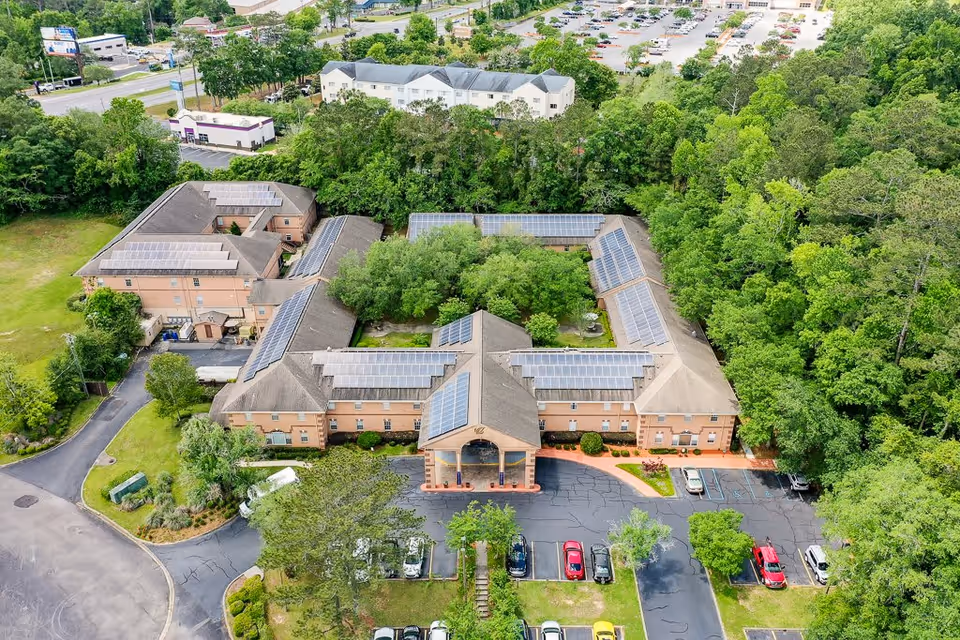 Aerial view of Woodmont Senior Living facility showing a large building with multiple wings arranged around a central courtyard with trees. The building has solar panels on the roofs and is surrounded by greenery and parking lots with several cars parked.