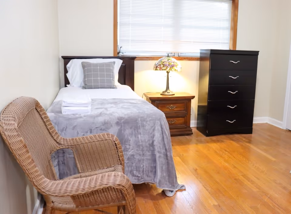 A compact bedroom in a senior facility featuring a bed, dresser, nightstand, lamp, and wicker chair.