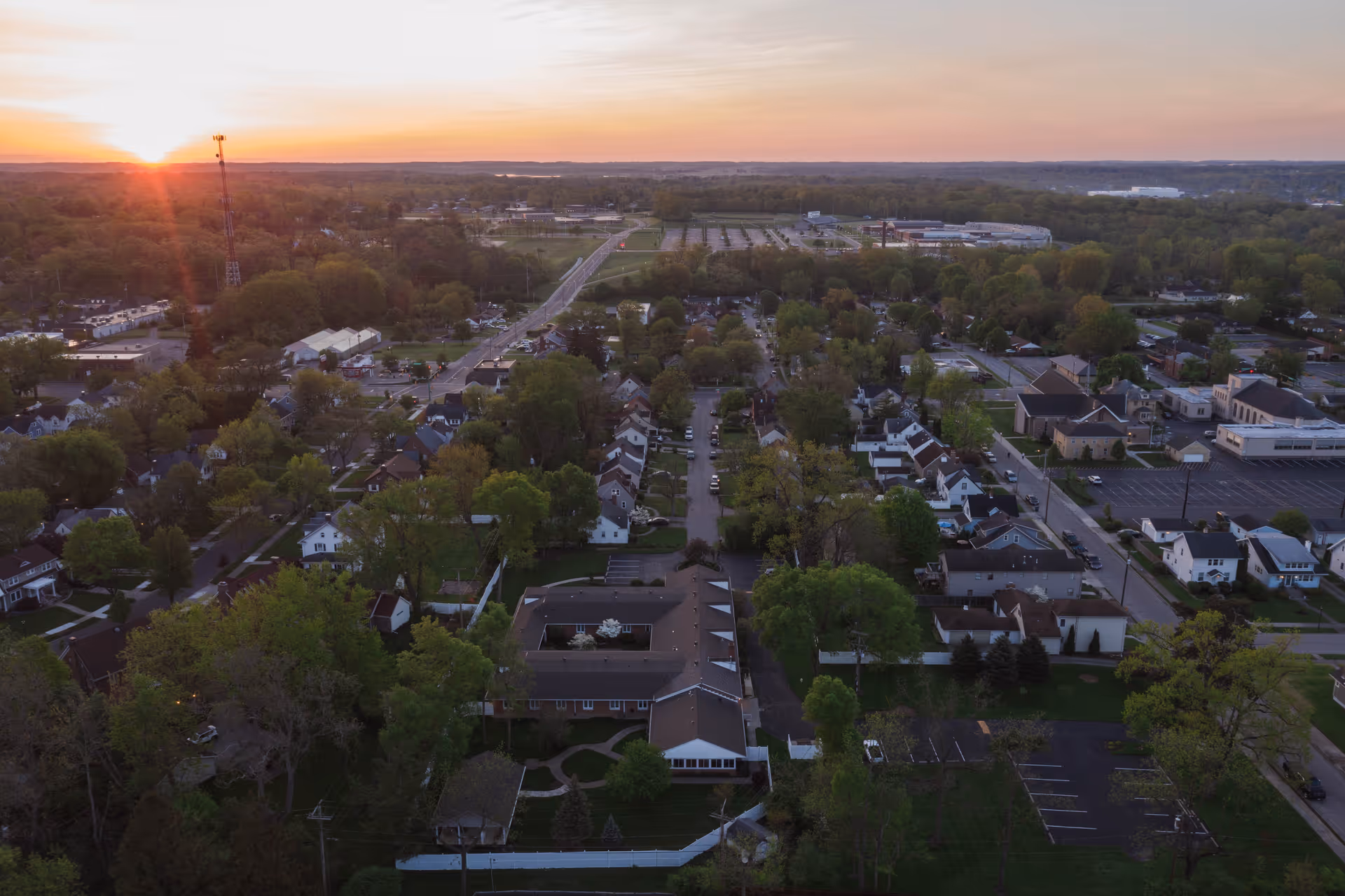 Aerial view of a suburban neighborhood at sunset with numerous houses, trees, roads, and parking lots visible, showing a peaceful residential area with the sun setting on the horizon.