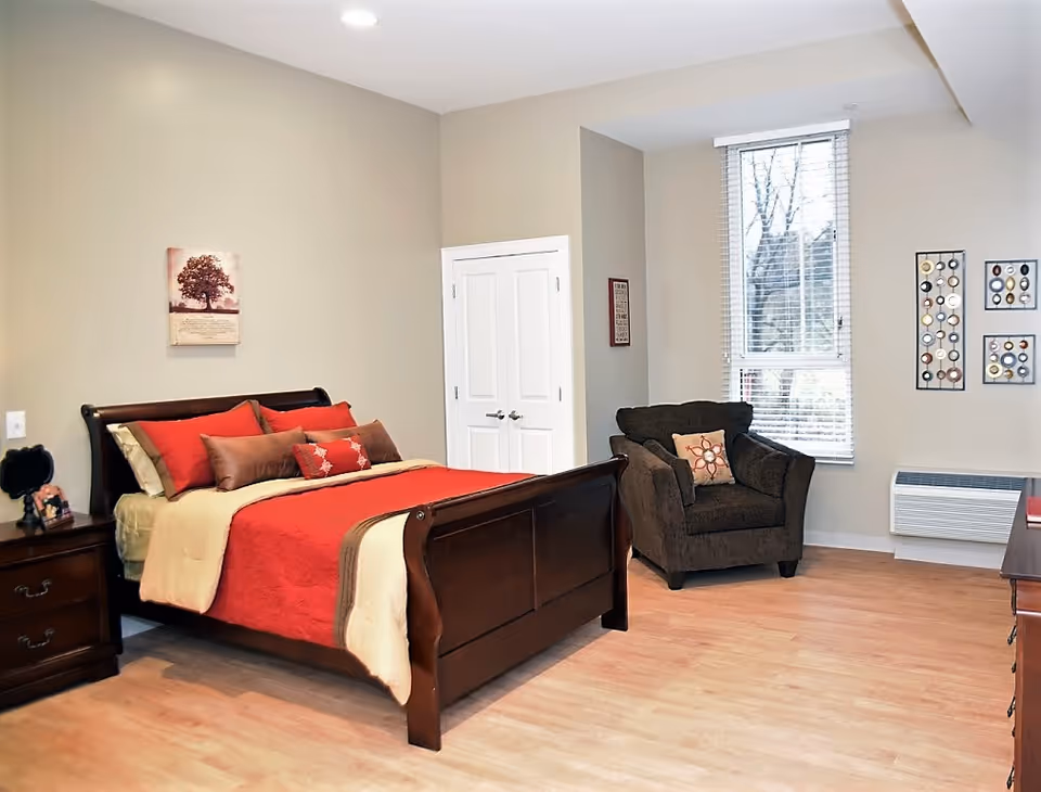 A furnished bedroom in a senior living facility with a bed, chair, and dresser.
