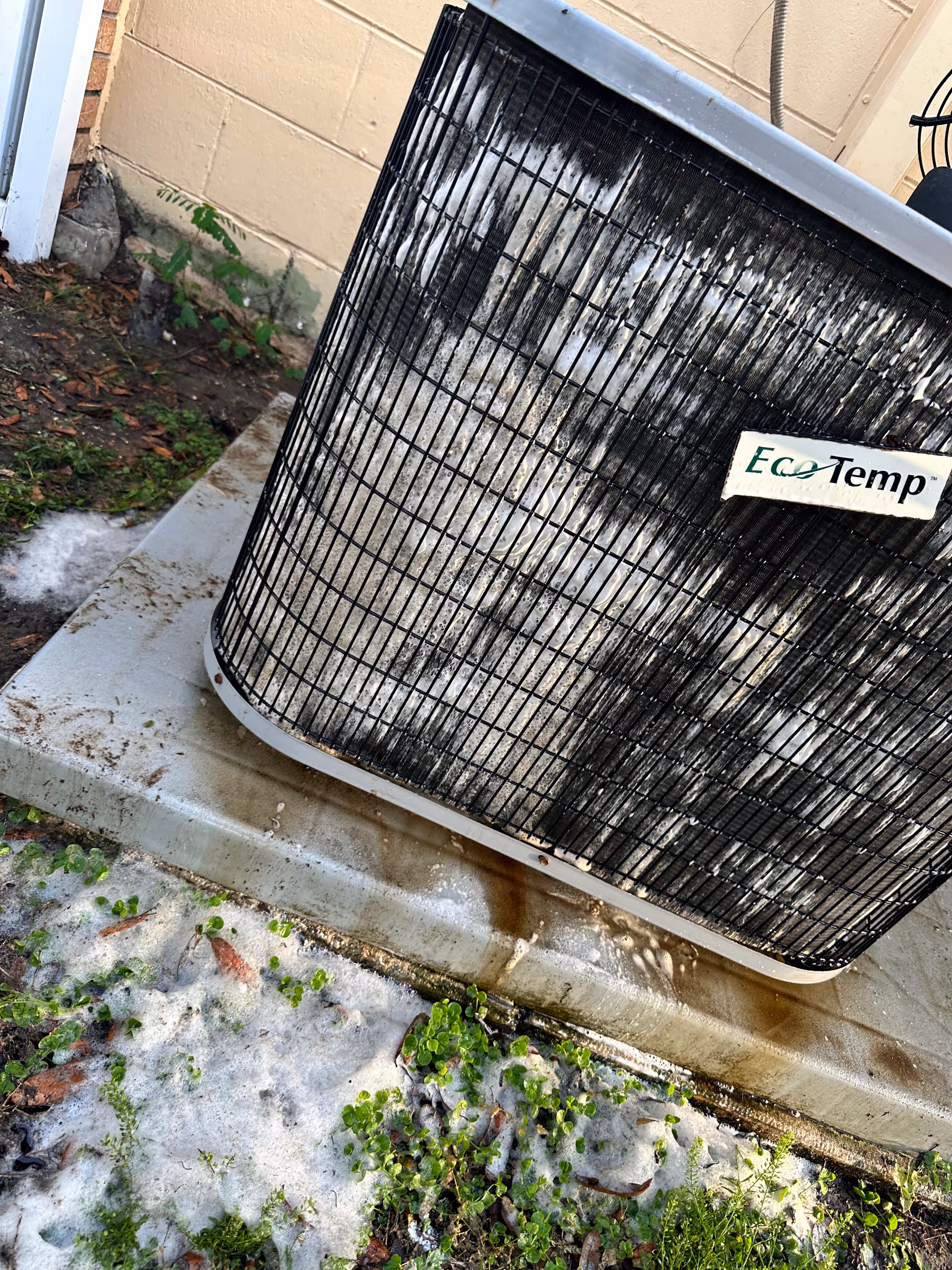 Outdoor air conditioning condenser sits on a concrete pad beside a building, with greenery nearby.