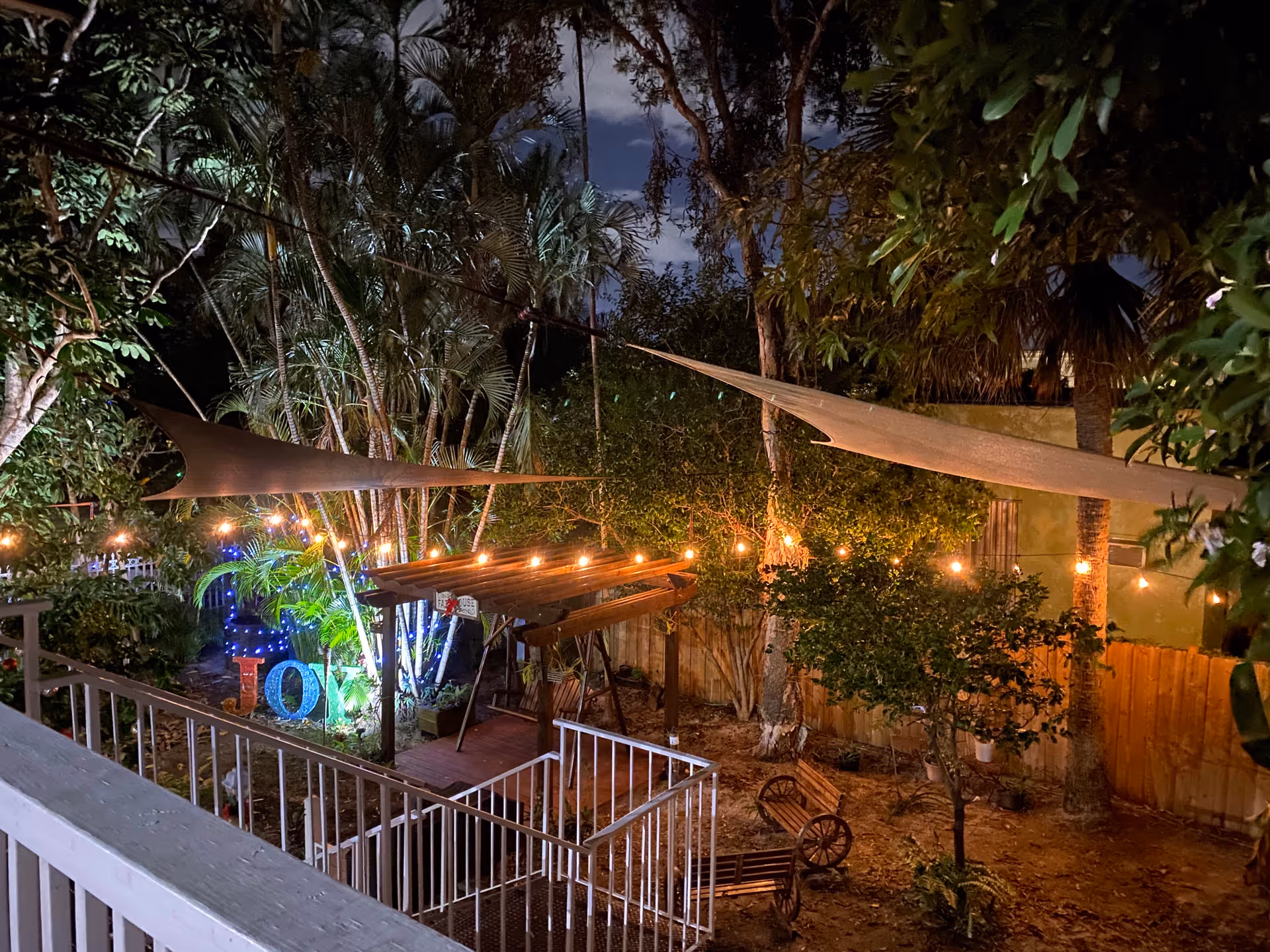 Nighttime view of a garden area with string lights hanging above, a wooden pergola, benches, and various trees and plants. The garden is enclosed by a wooden fence and has decorative lighting spelling out the word 'LOVE'.