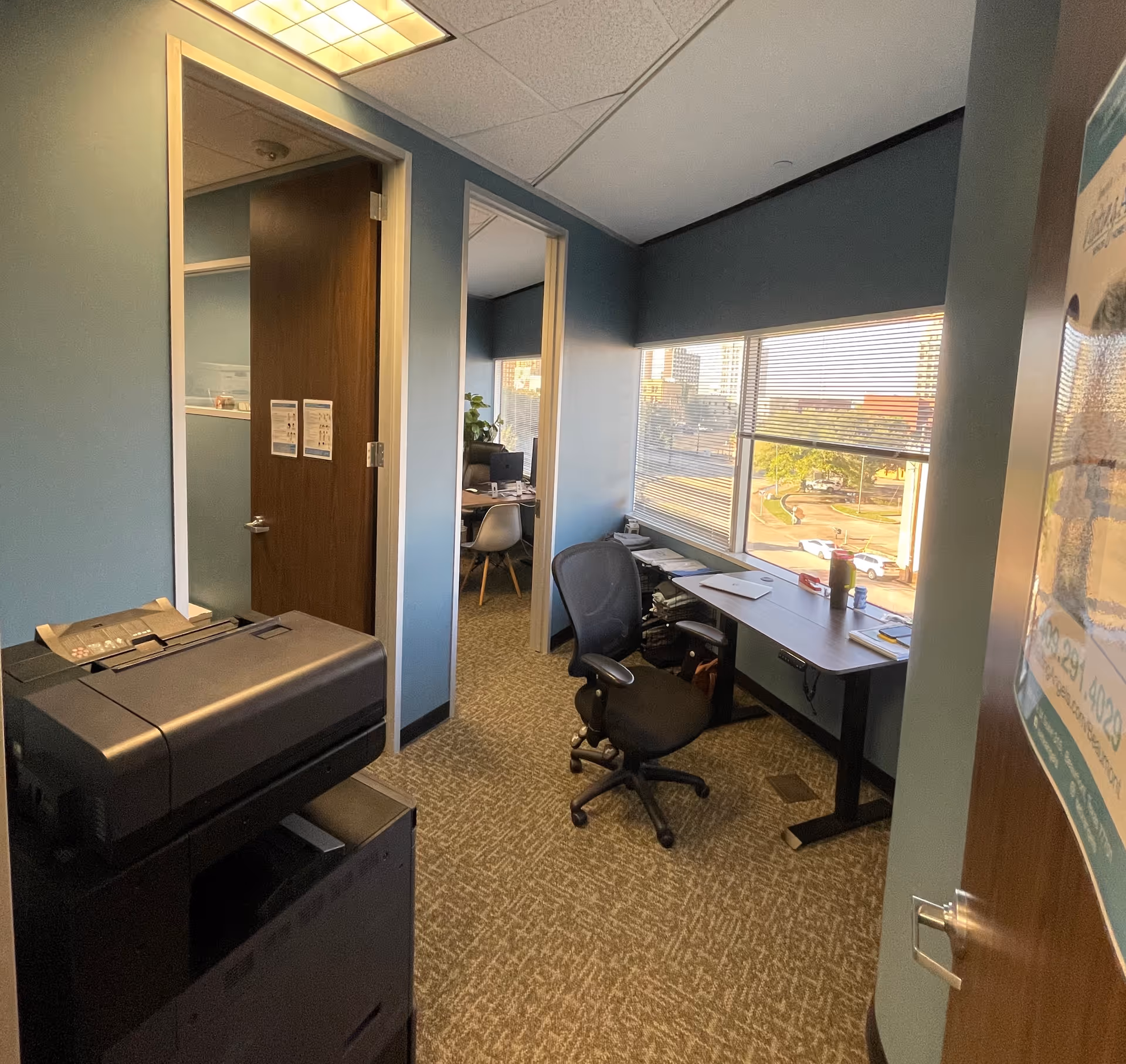 Office workspace in a blue-walled senior facility with desk, chair, printer, and window view.