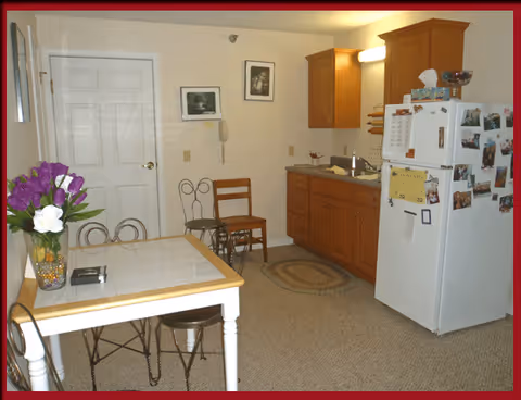 Small kitchenette and dining area with a table and chairs, a white refrigerator, wooden cabinets, and a vase of flowers on the table.