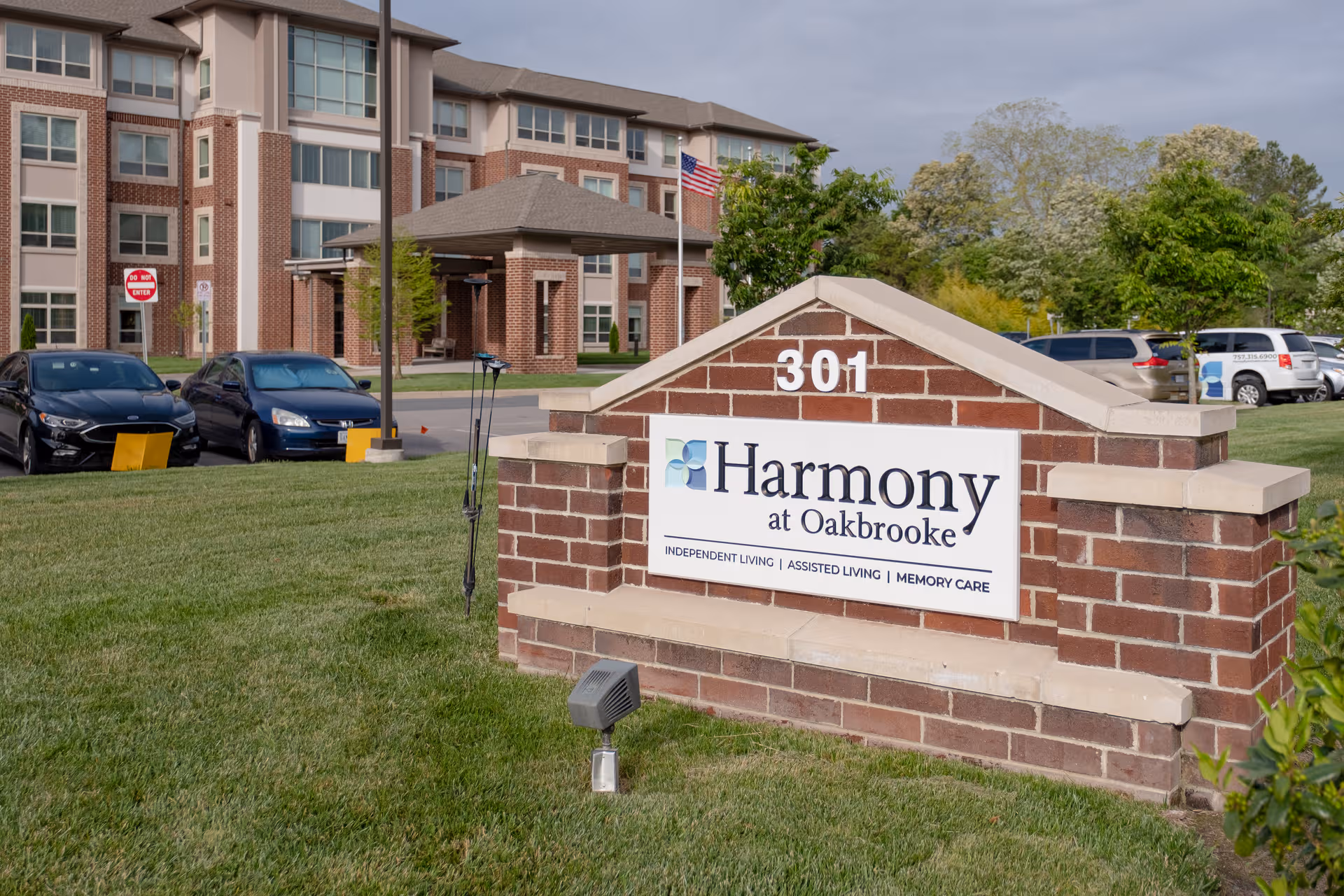 Brick entrance sign reading Harmony at Oakbrooke with a brick building and parked cars in view.
