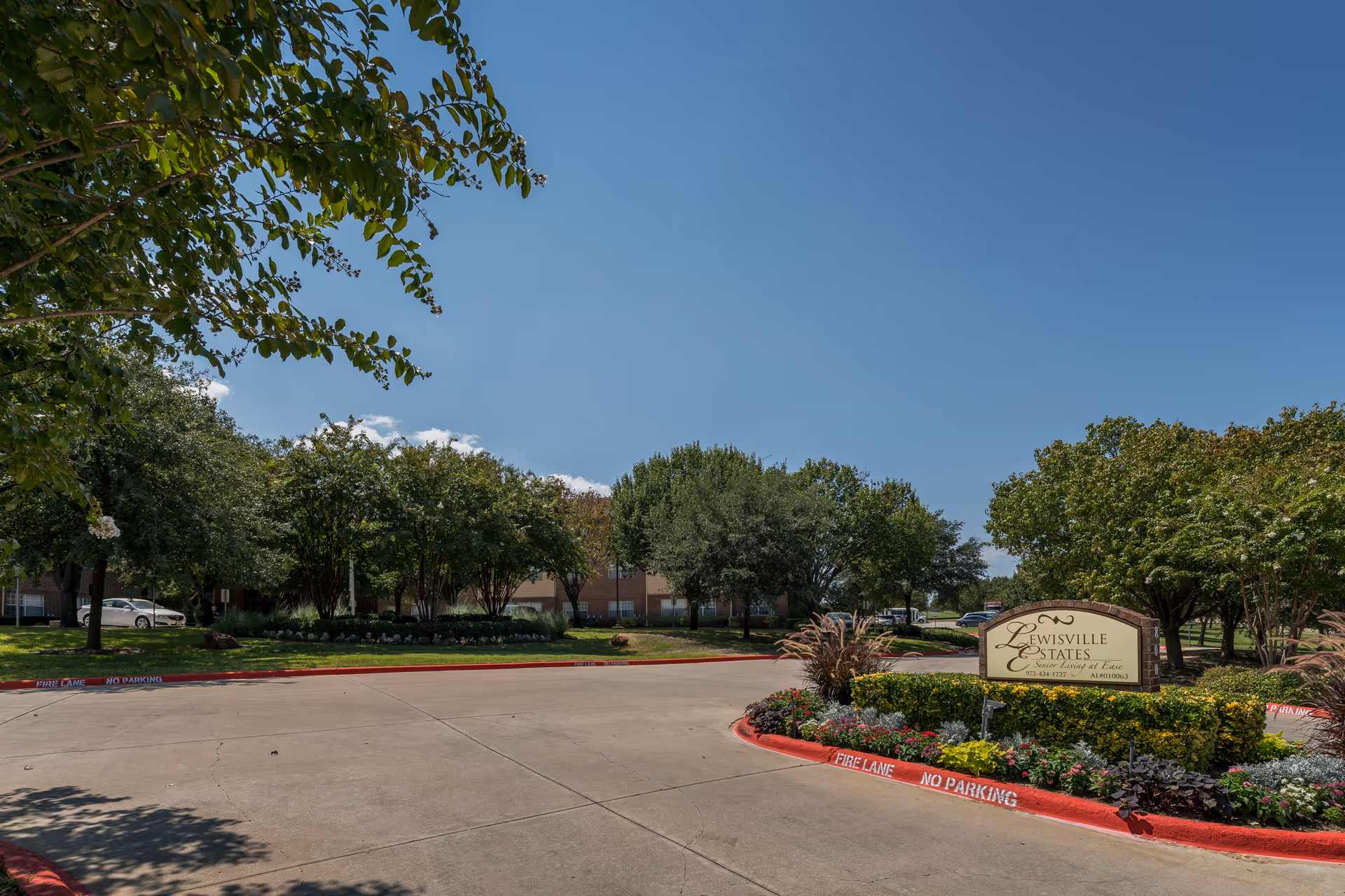 Front exterior of Lewisville Estates with curved driveway, landscaped beds, and a sign.