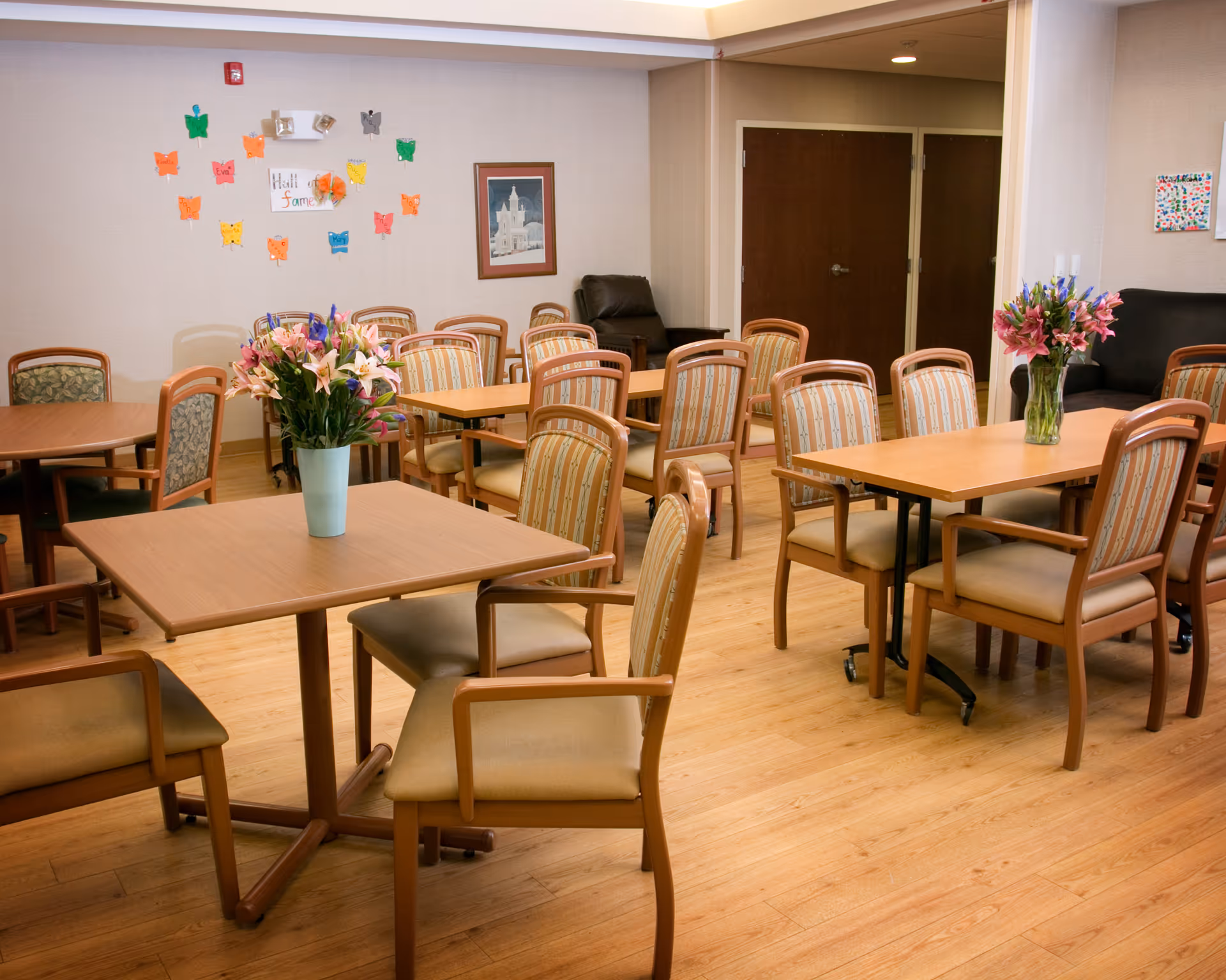 Bright dining room with multiple tables, chairs, and colorful flower arrangements.
