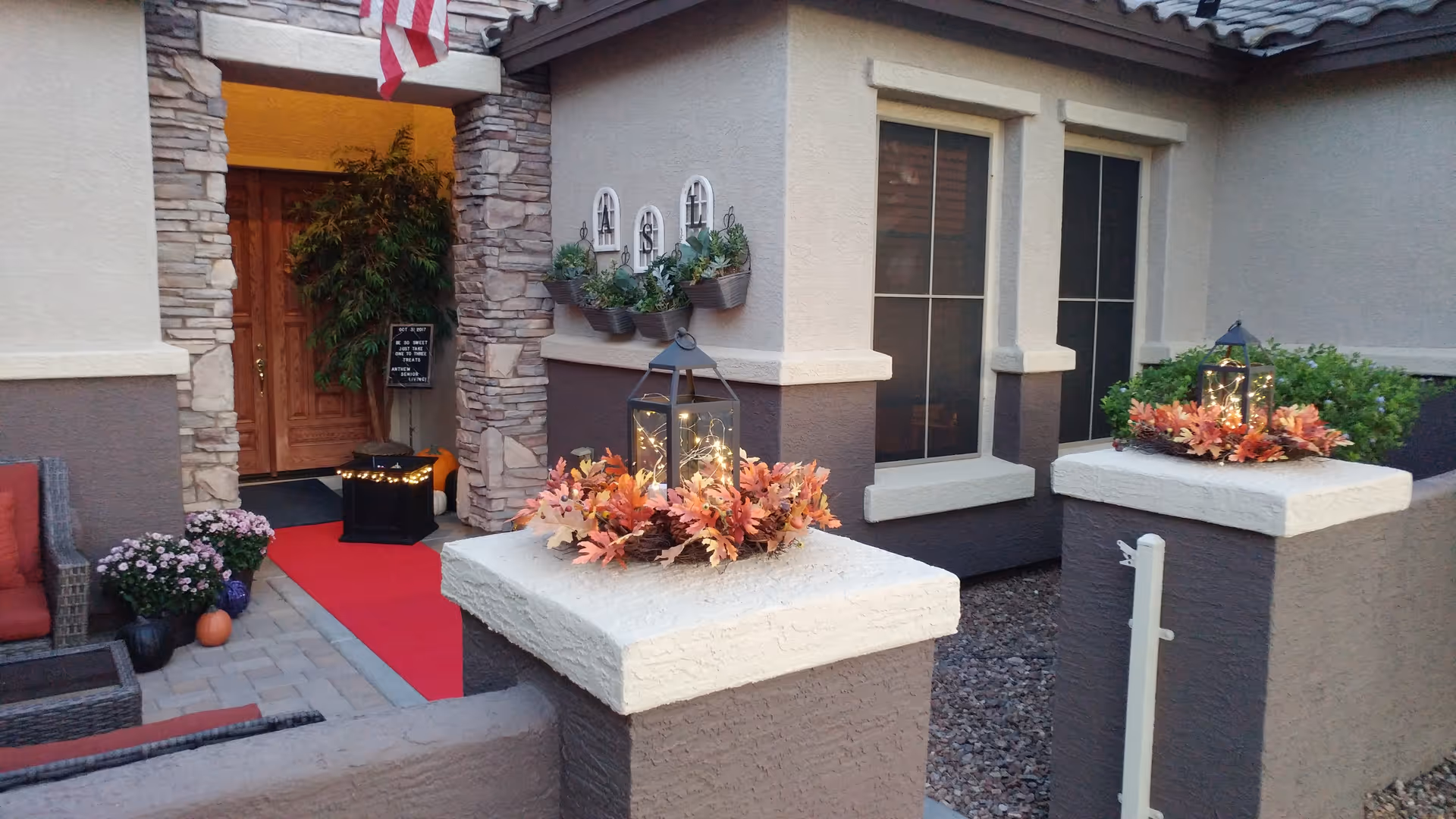 Front entrance of a home with stone pillars, a red runner, lanterns and autumn floral decorations on low walls.
