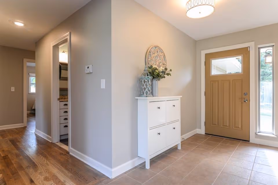Entryway of a residence with a tan front door featuring a window panel, a narrow vertical window to the right, and a white cabinet with decorative items on top against a beige wall. Hardwood flooring leads down a hallway with a partially visible room and bathroom on the left.