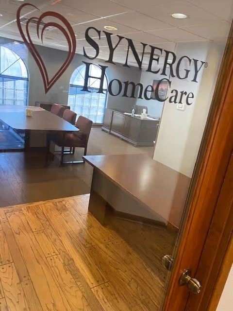 Glass wall displays SYNERGY HomeCare logo; interior office with table and chairs.