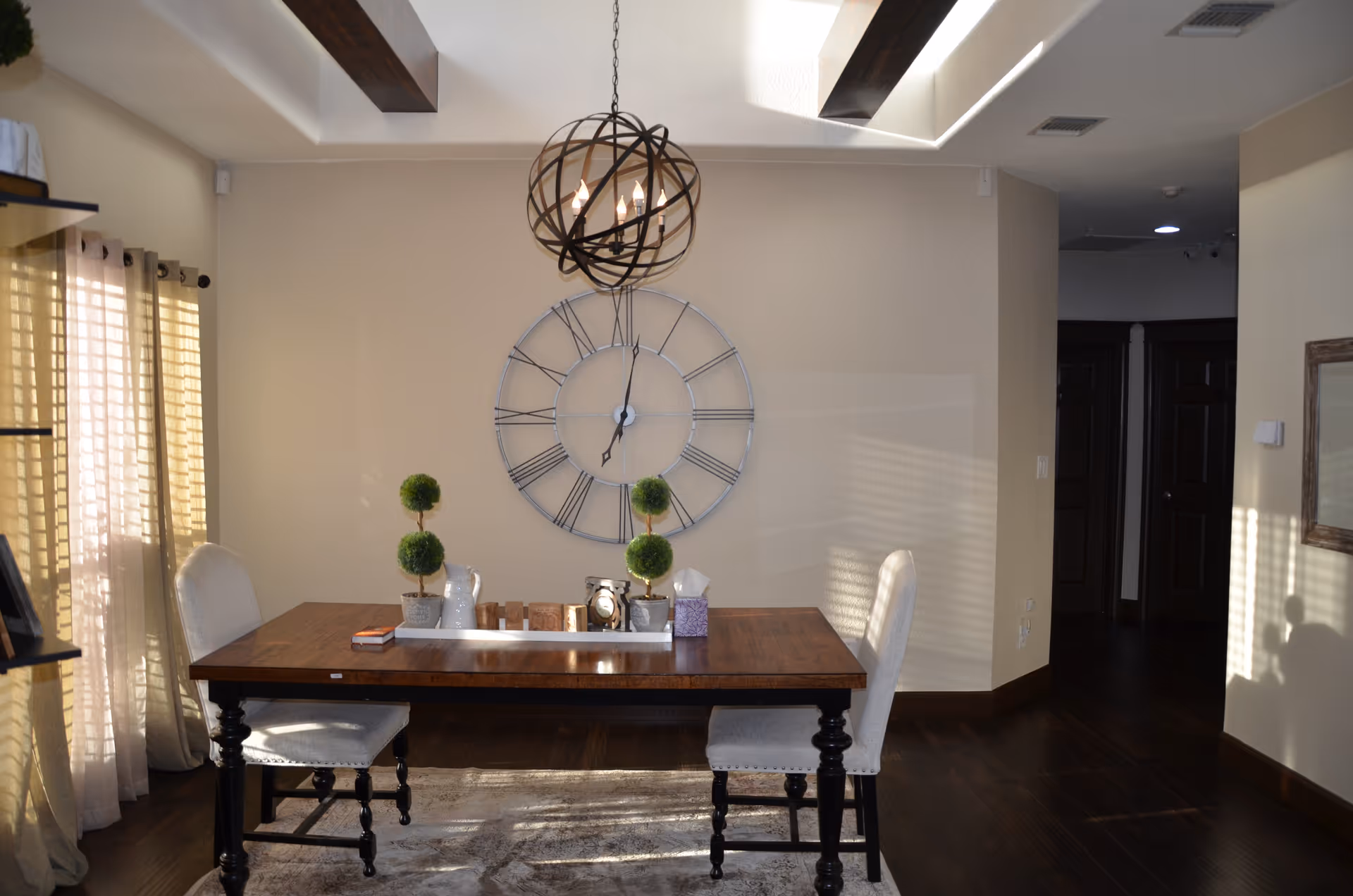 A dining area with a wooden table and two upholstered chairs. The table has decorative items including small potted plants, a tissue box, and a clock. A large wall clock with Roman numerals hangs on the beige wall behind the table. A modern spherical chandelier hangs from the ceiling, and sunlight filters through sheer curtains on the left side.