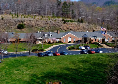Front exterior of a single-story brick senior living building with a circular driveway, parked cars, flagpole, and a large lawn.