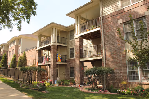 Exterior view of a multi-story senior living facility building with brick and siding facade, balconies, a fenced garden area, and a sidewalk in front.