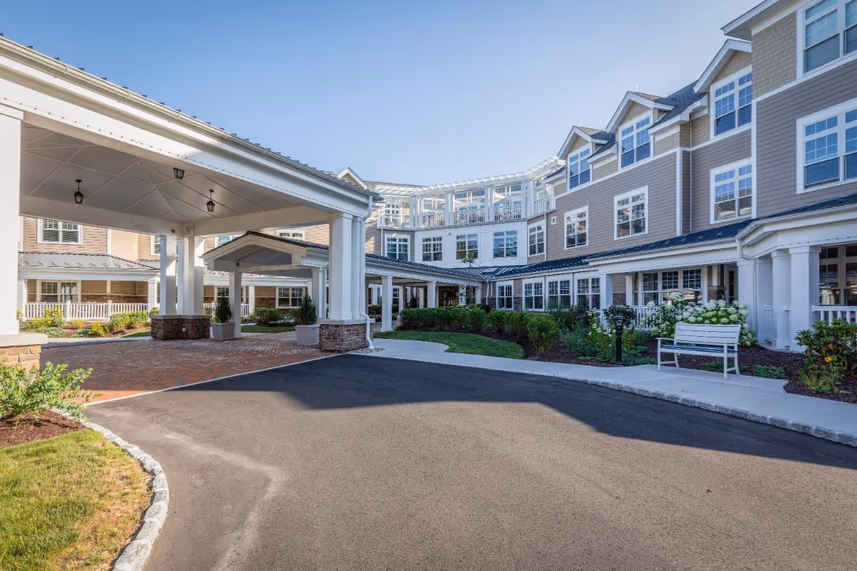 Front entrance of a multi-story senior living building with a covered porte-cochere, driveway, benches, and landscaped grounds.