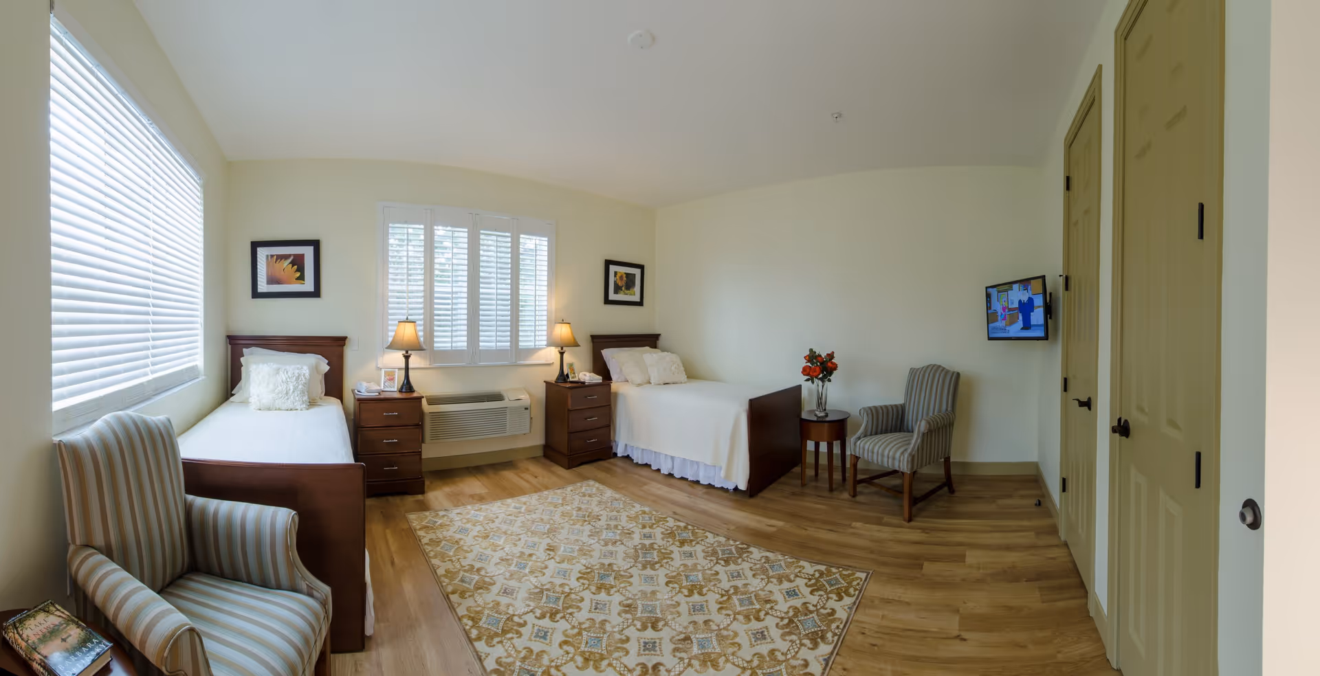 A bright, clean bedroom with two single beds, each with white bedding and decorative pillows. Between the beds are two wooden nightstands with lamps and a telephone. The room has light-colored walls, a large window with blinds, two framed pictures of sunflowers, a patterned area rug, two striped armchairs, a small round table with a vase of flowers, and a wall-mounted TV.