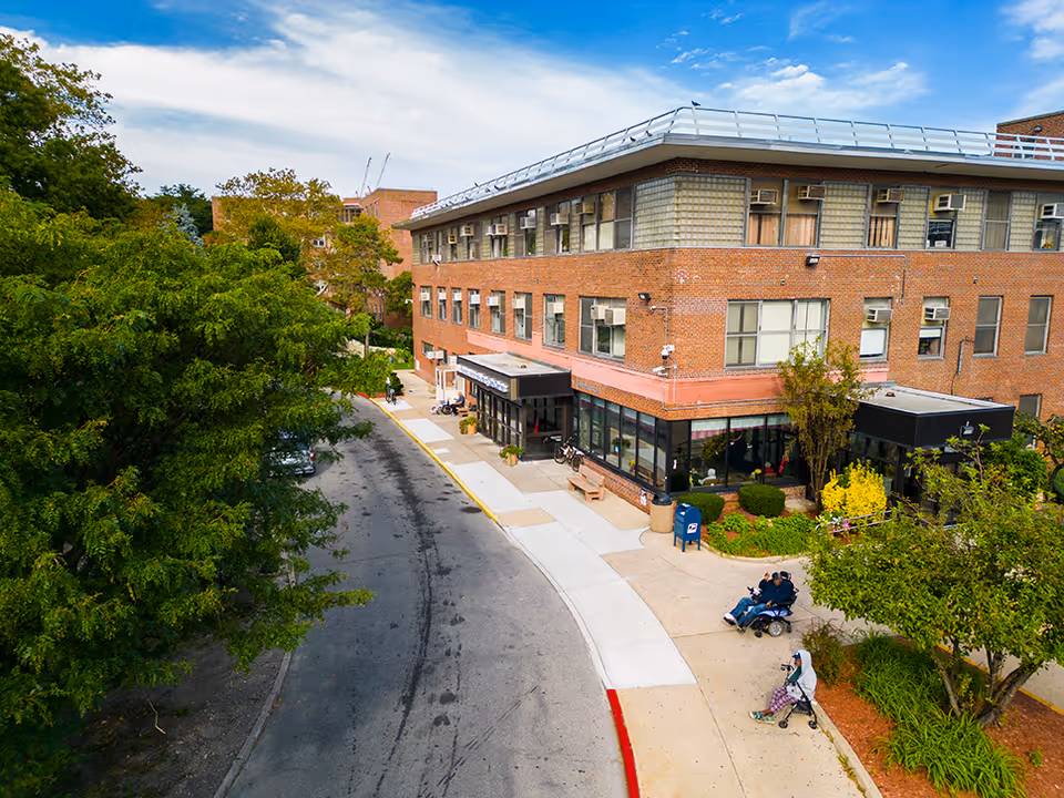 A brick senior living facility exterior with storefronts, sidewalk, and surrounding trees.