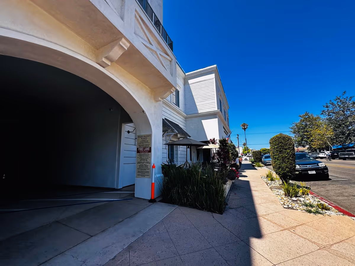 Street-level view of the Monarch Cottages building entrance and sidewalk with parked cars and landscaping under a clear blue sky.