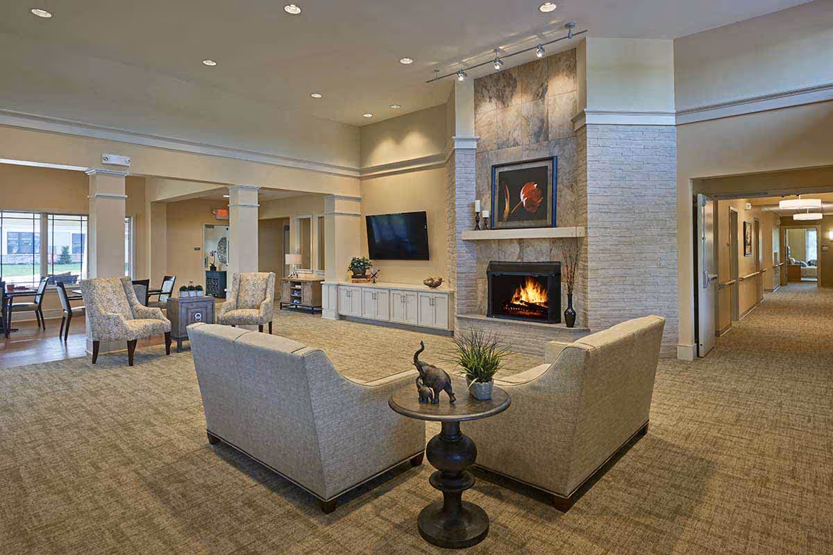 A spacious and well-lit living room area in a senior living facility with beige carpeting and neutral-colored walls. The room features a stone fireplace with a fire burning, a framed floral artwork above it, and a flat-screen TV mounted on the wall. There are two beige sofas arranged around a small round table with decorative items, and two patterned armchairs near a small square table. In the background, there is a dining area with tables and chairs near large windows, and a hallway leading to other rooms.
