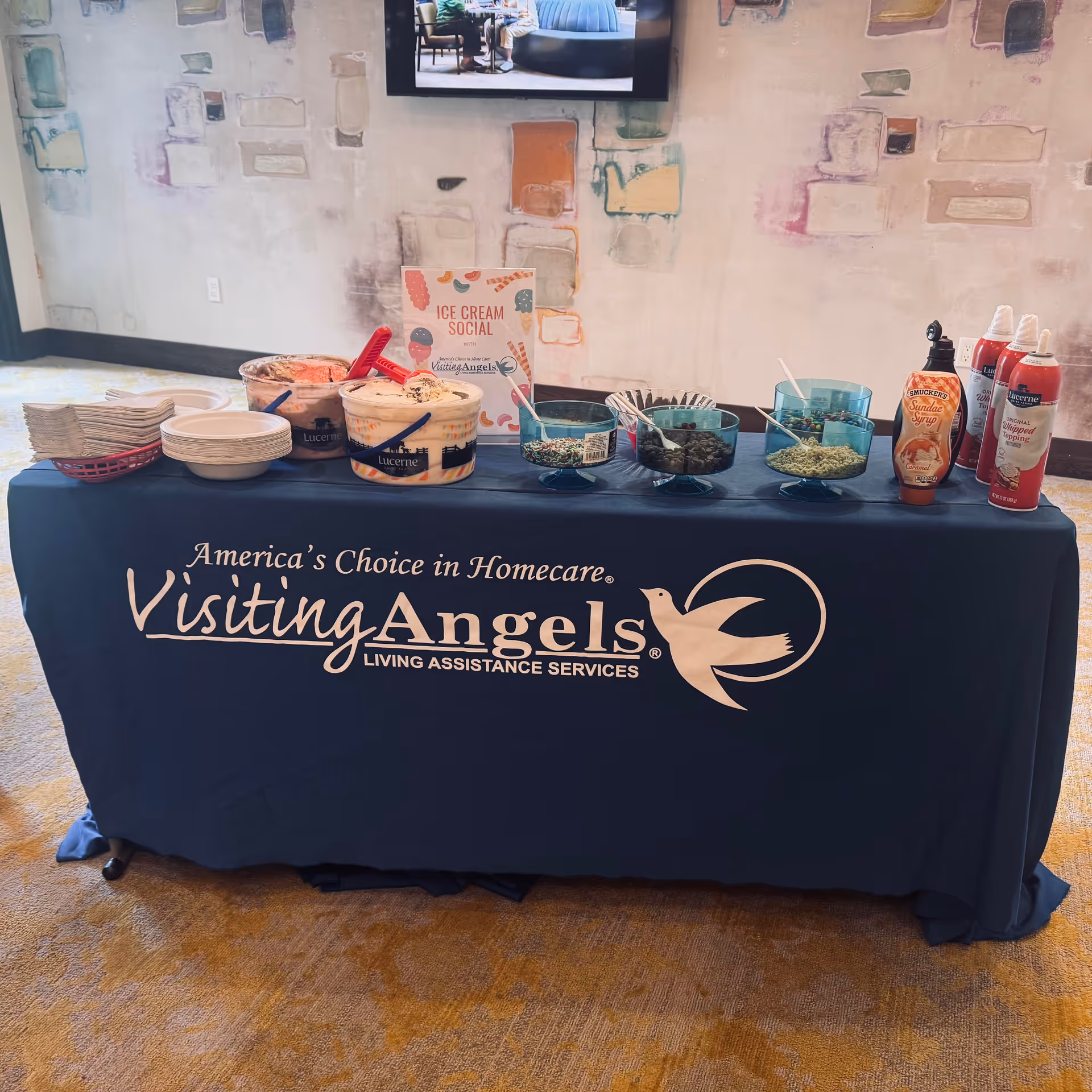 A navy tablecloth table with ice cream toppings, sauces, and a Visiting Angels banner.