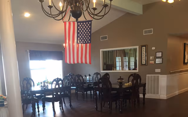 Dining room with dark wooden tables and chairs, American flag, chandelier, and windows.