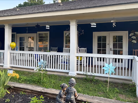 Blue exterior building with a covered porch, white columns, seating, and garden statues in front.