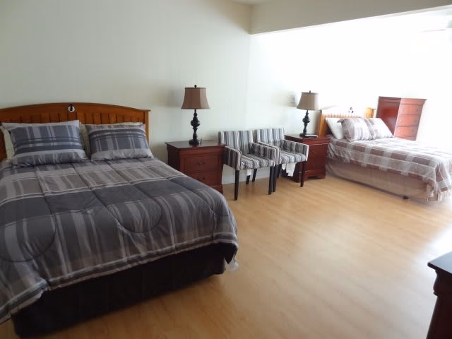 Spacious bedroom with two beds dressed in matching plaid bedding, nightstands with lamps, two striped chairs, and light wood flooring.