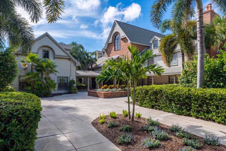 Exterior view of a senior living facility named South Port Square featuring a paved walkway, landscaped garden beds with small palm trees and shrubs, and a multi-story building with a mix of brick and siding under a partly cloudy blue sky.
