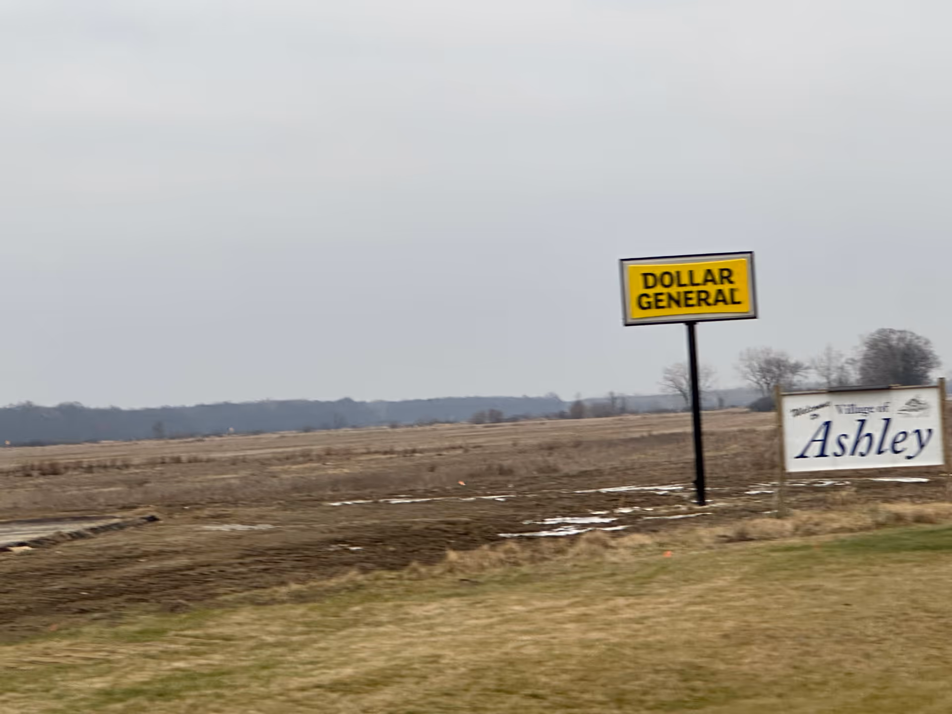 Wide rural scene with a Dollar General sign and an Ashley village sign.