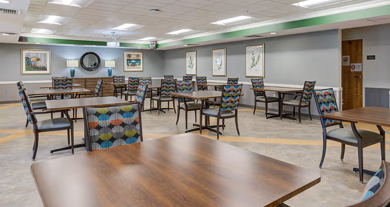 A spacious dining room with multiple wooden tables and chairs featuring colorful patterned upholstery. The room has framed artwork and botanical prints on the walls, a round mirror, and soft lighting from wall sconces. The floor is a light-colored tile with a subtle pattern, and the ceiling has recessed lighting and a projector.