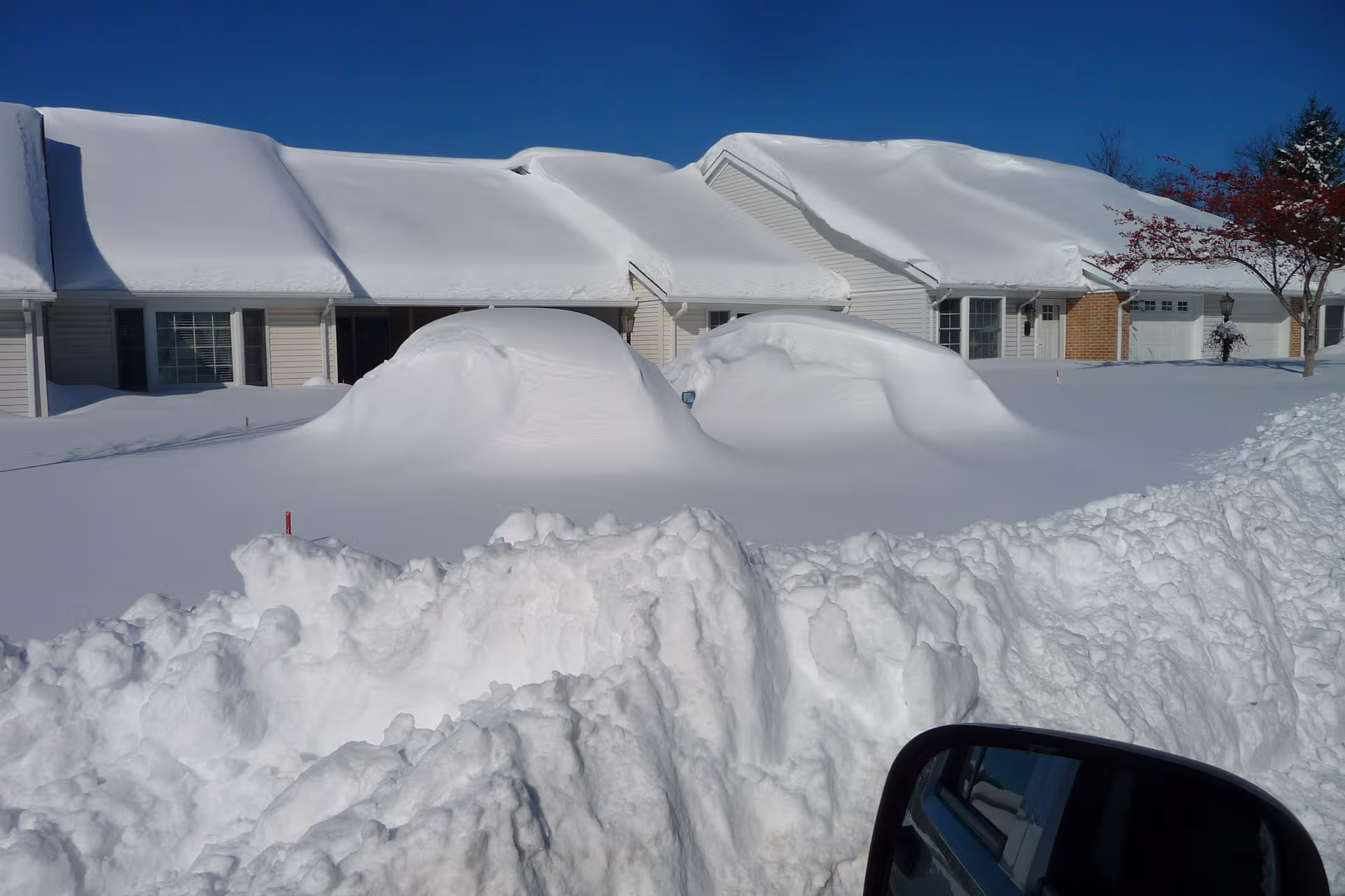 Snow covers the front of a senior living facility, with heavy snowbanks and a car mirror.