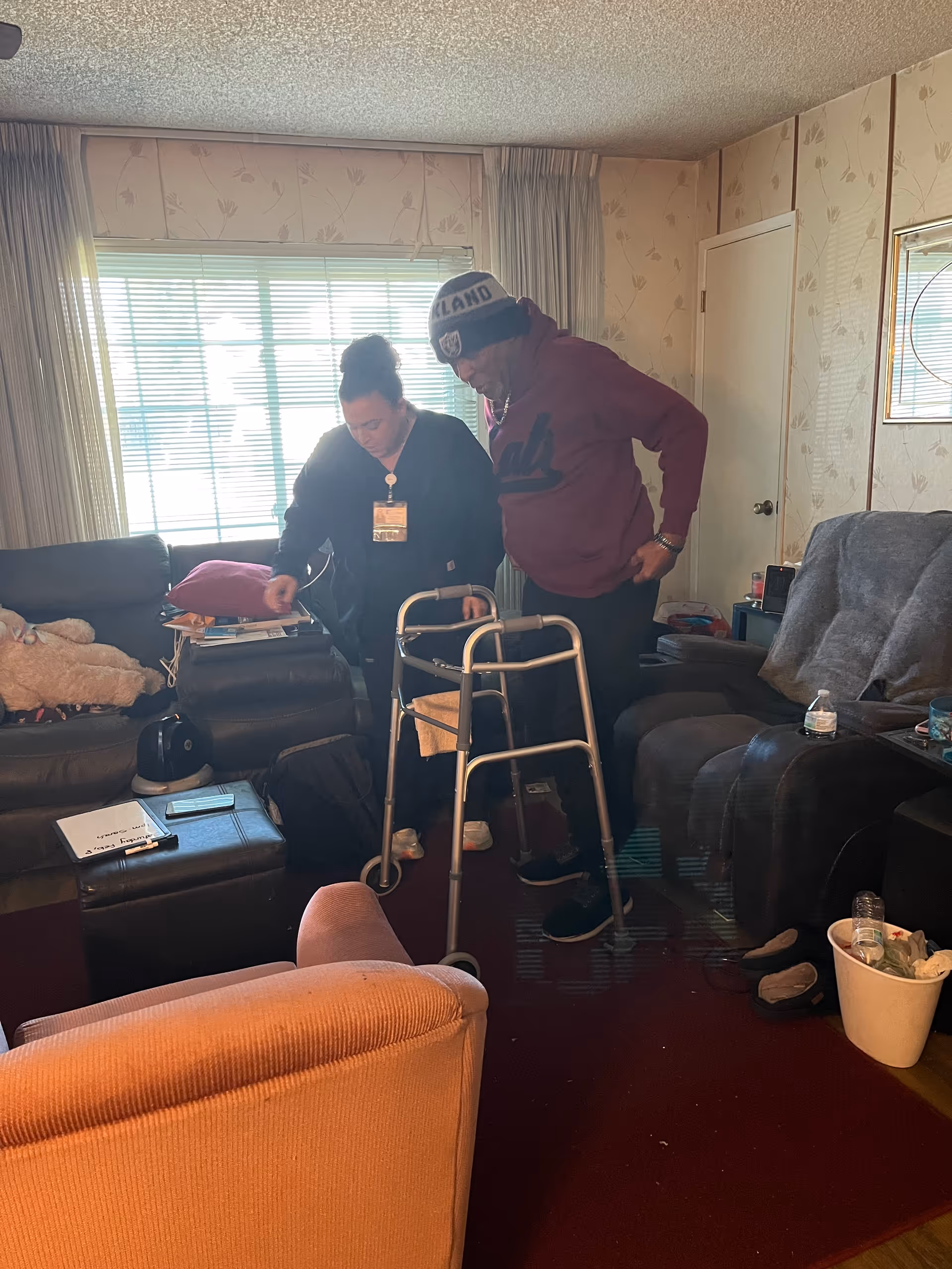Two adults stand near a walker in a cozy, cluttered living room.