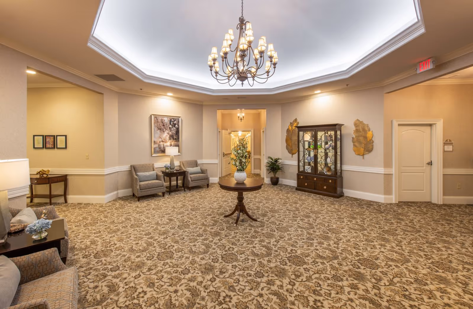 A spacious, well-lit common area with patterned carpet, beige walls, and a large chandelier hanging from a recessed ceiling. The room features a round wooden table with a floral arrangement in the center, several upholstered chairs, a wooden display cabinet with decorative items, and framed artwork on the walls. There are doorways leading to other rooms and an exit sign above one door.