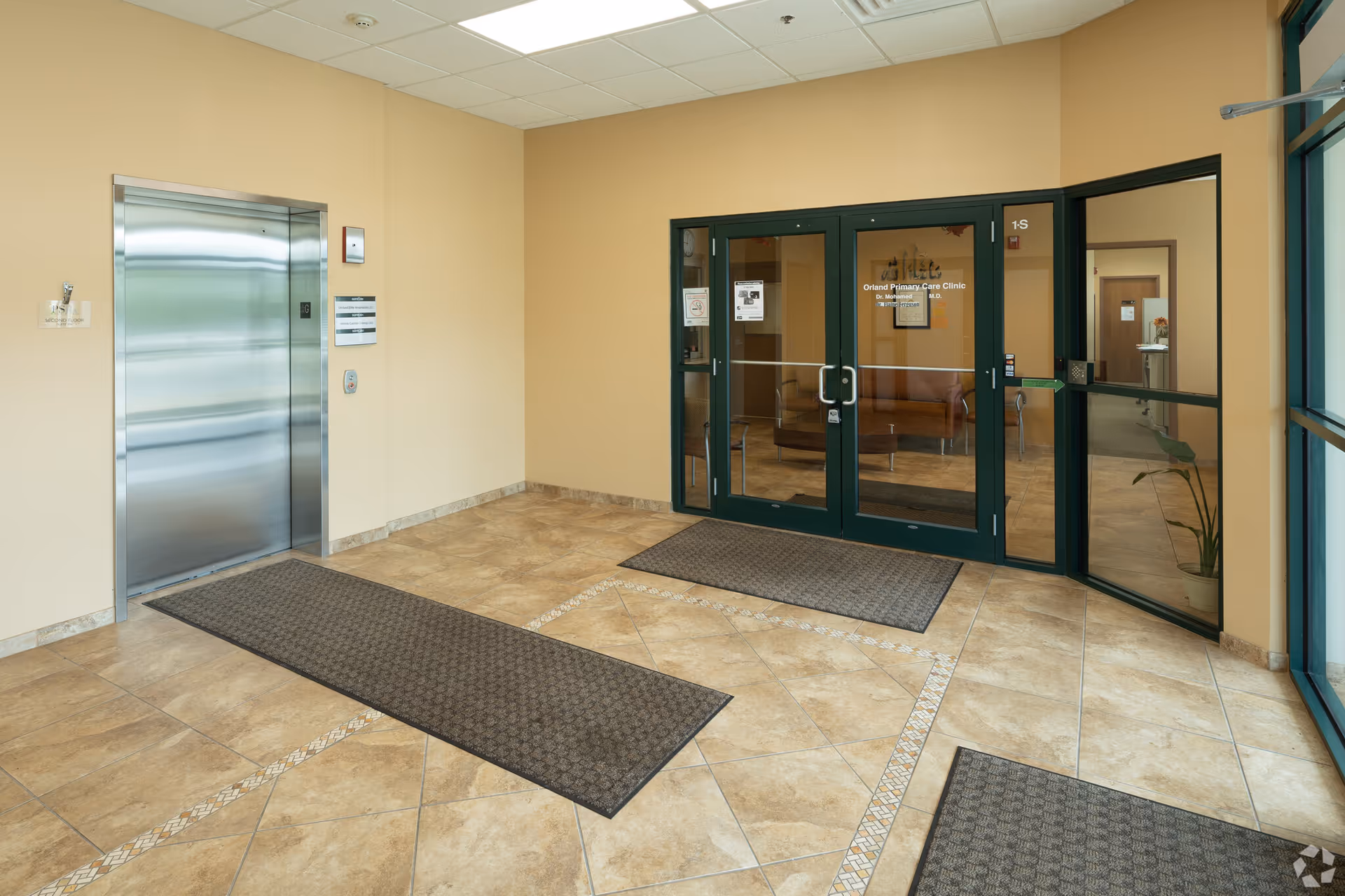 Lobby of a senior facility with glass doors, elevator, beige walls, and tiled floor.