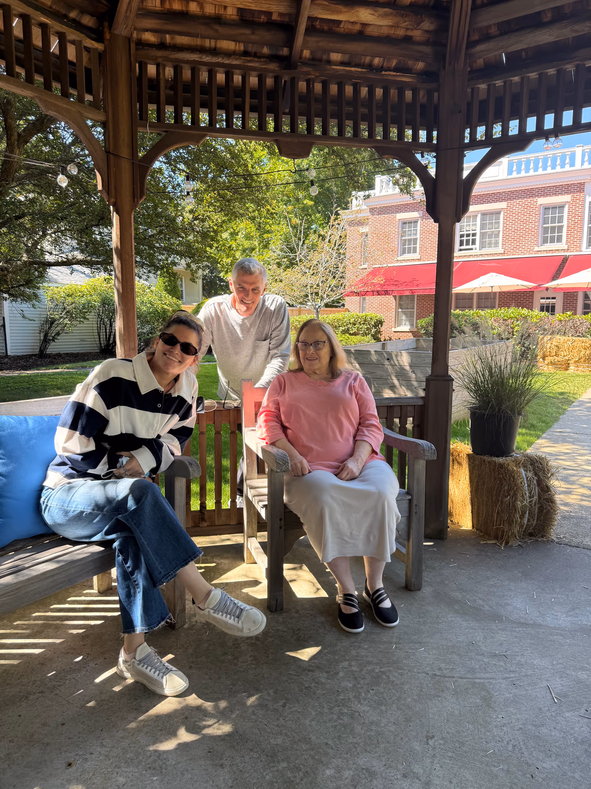 Three people sit on wooden benches in a gazebo outdoors with a brick building behind.