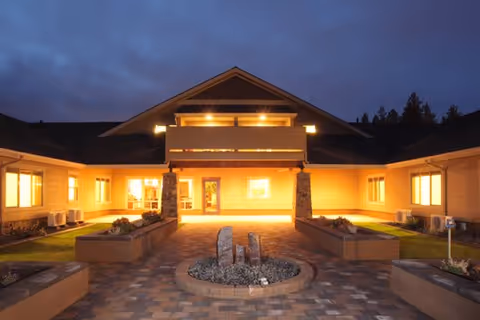 Exterior view of a single-story building at dusk with warm lights glowing from the windows and entrance. The building has a covered entryway supported by stone pillars and a paved walkway leading to the door, with landscaped planters on either side.