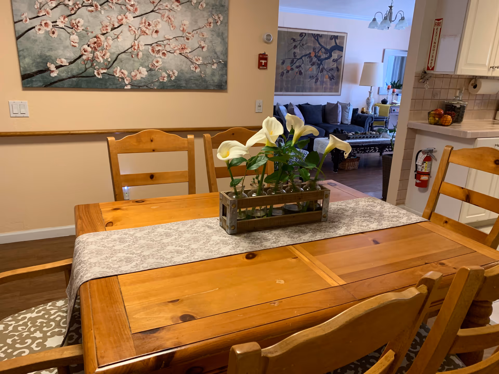 Wood dining table with floral centerpiece and chairs, in a dining area near the adjacent kitchen.