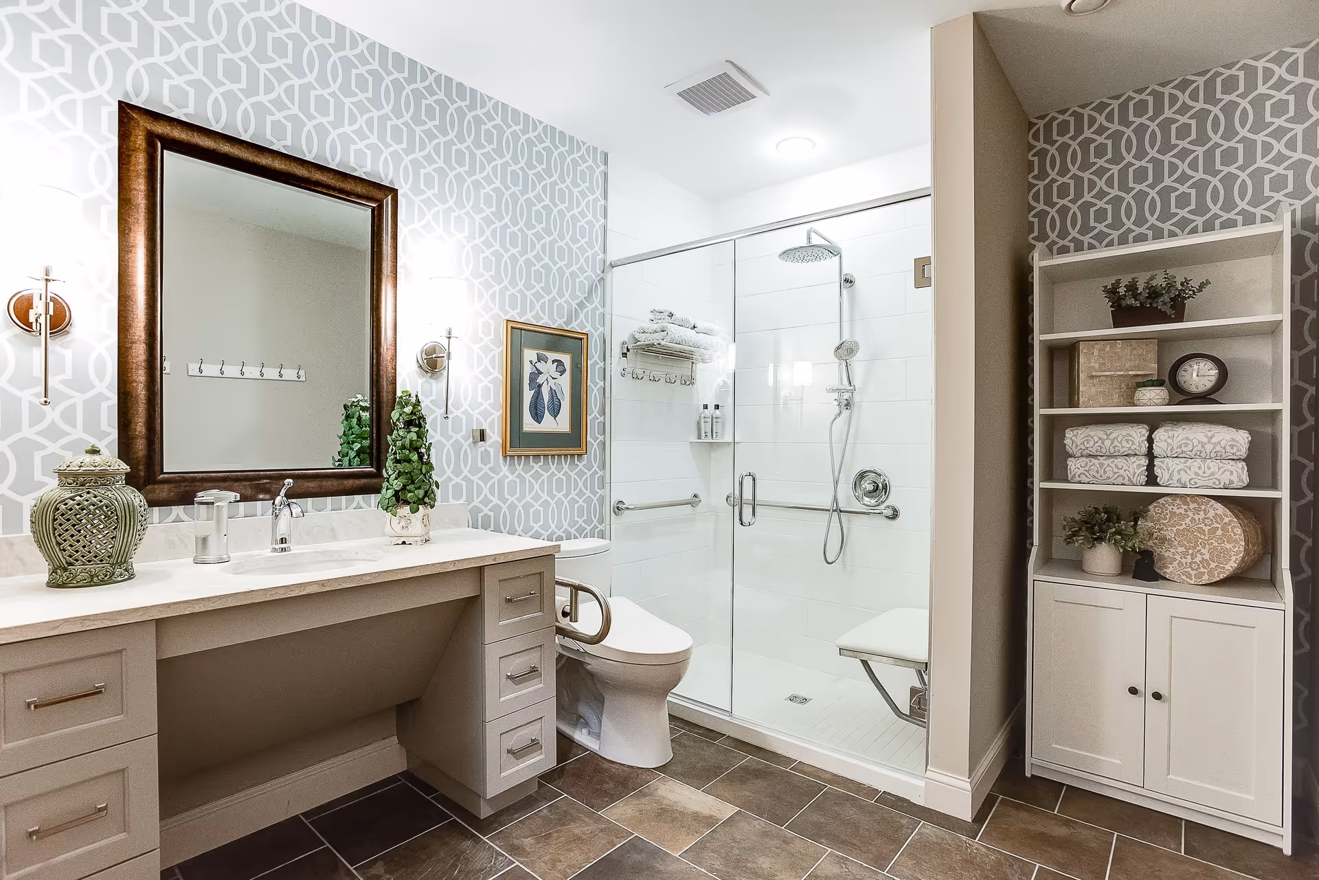 A modern bathroom with a large mirror above a vanity with a sink and decorative items. To the right is a toilet with a safety grab bar and a glass-enclosed walk-in shower featuring a shower seat, grab bars, and a rainfall showerhead. On the far right is a white shelving unit holding folded towels, plants, a clock, and decorative boxes. The walls have a geometric patterned wallpaper, and the floor is tiled in brown tones.