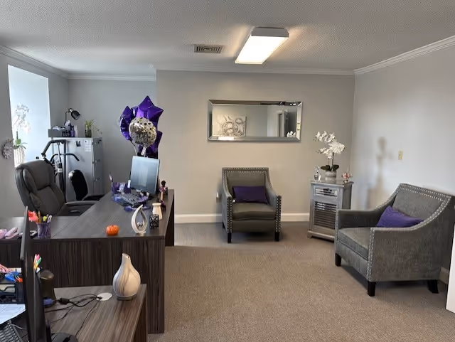 Office-style reception lounge with desk, two chairs, plants, and wall mirror in senior facility.
