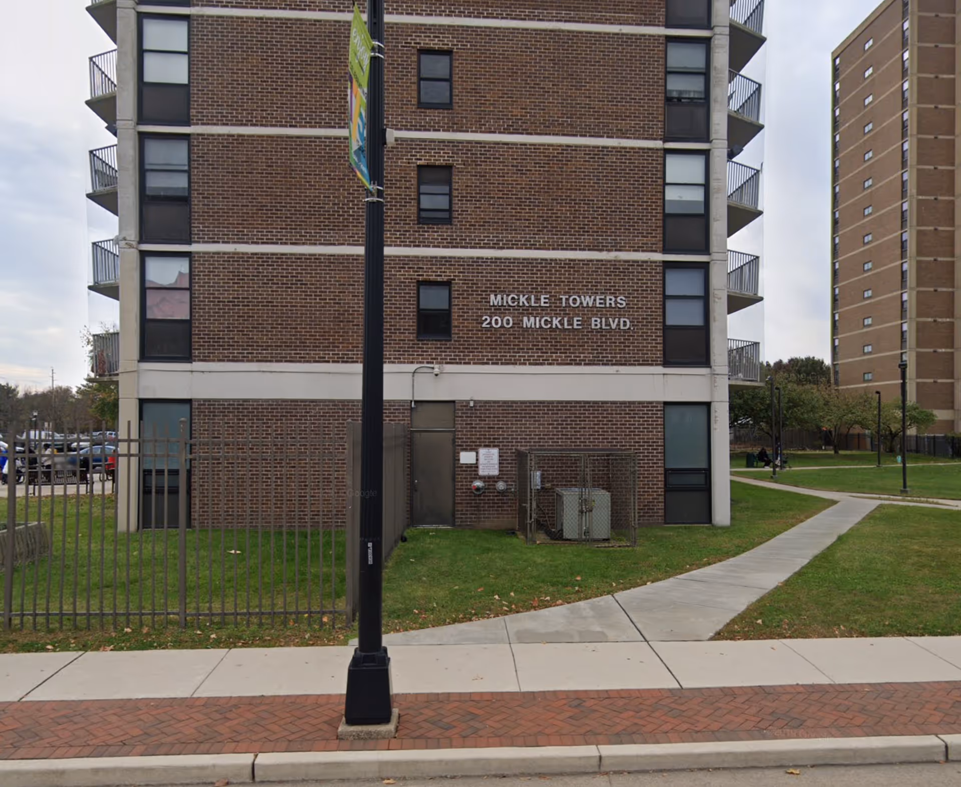 A brick mid-rise Mickle Towers building with sidewalk, lawn, and a fenced utility enclosure.