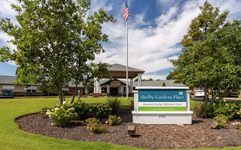 Front entrance of Shelby Gardens Place assisted living facility with a sign, flagpole, lawn, and landscaping.