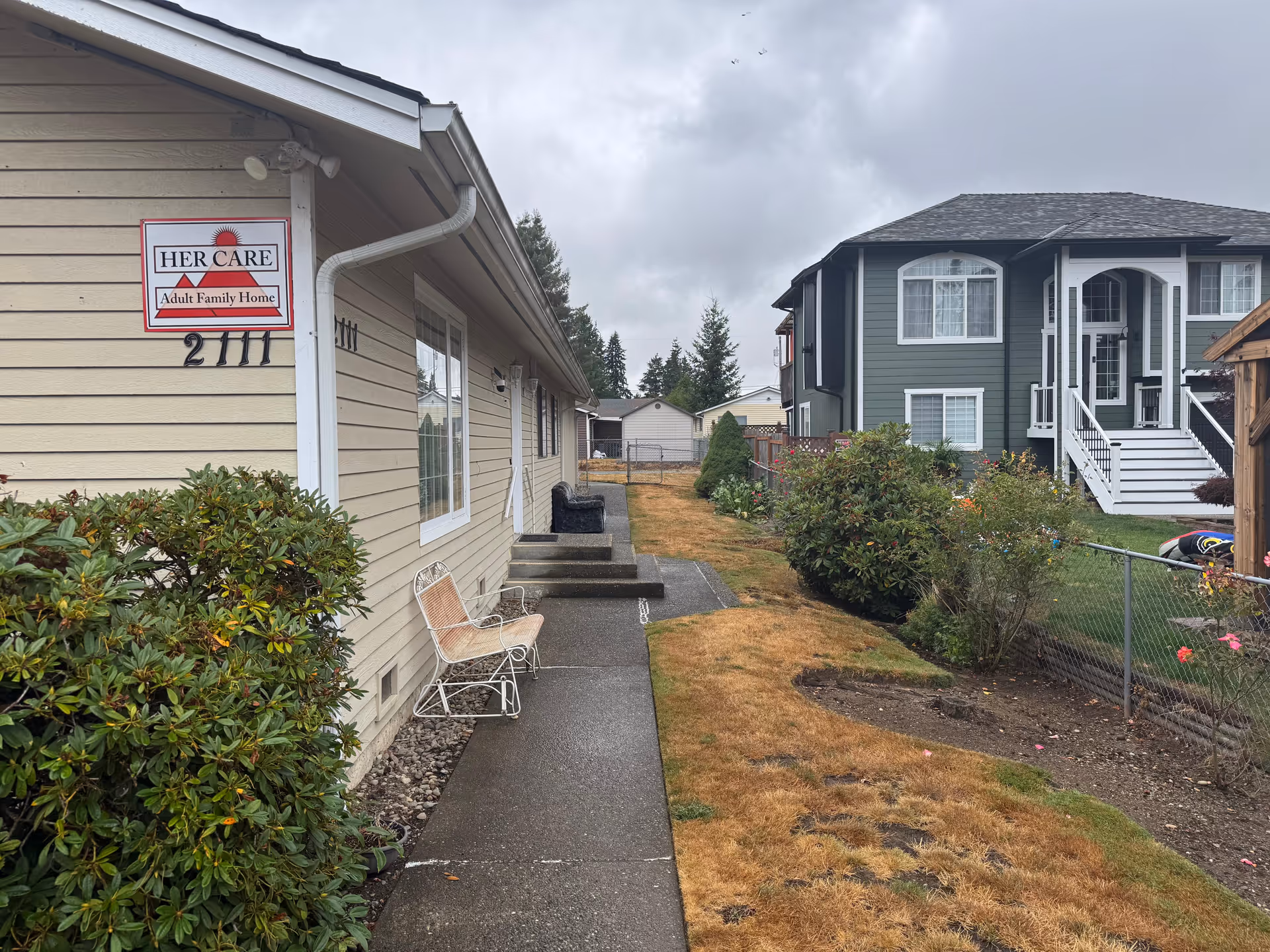 Exterior view of HER CARE Adult Family Home with walkway, chairs, and neighboring house.