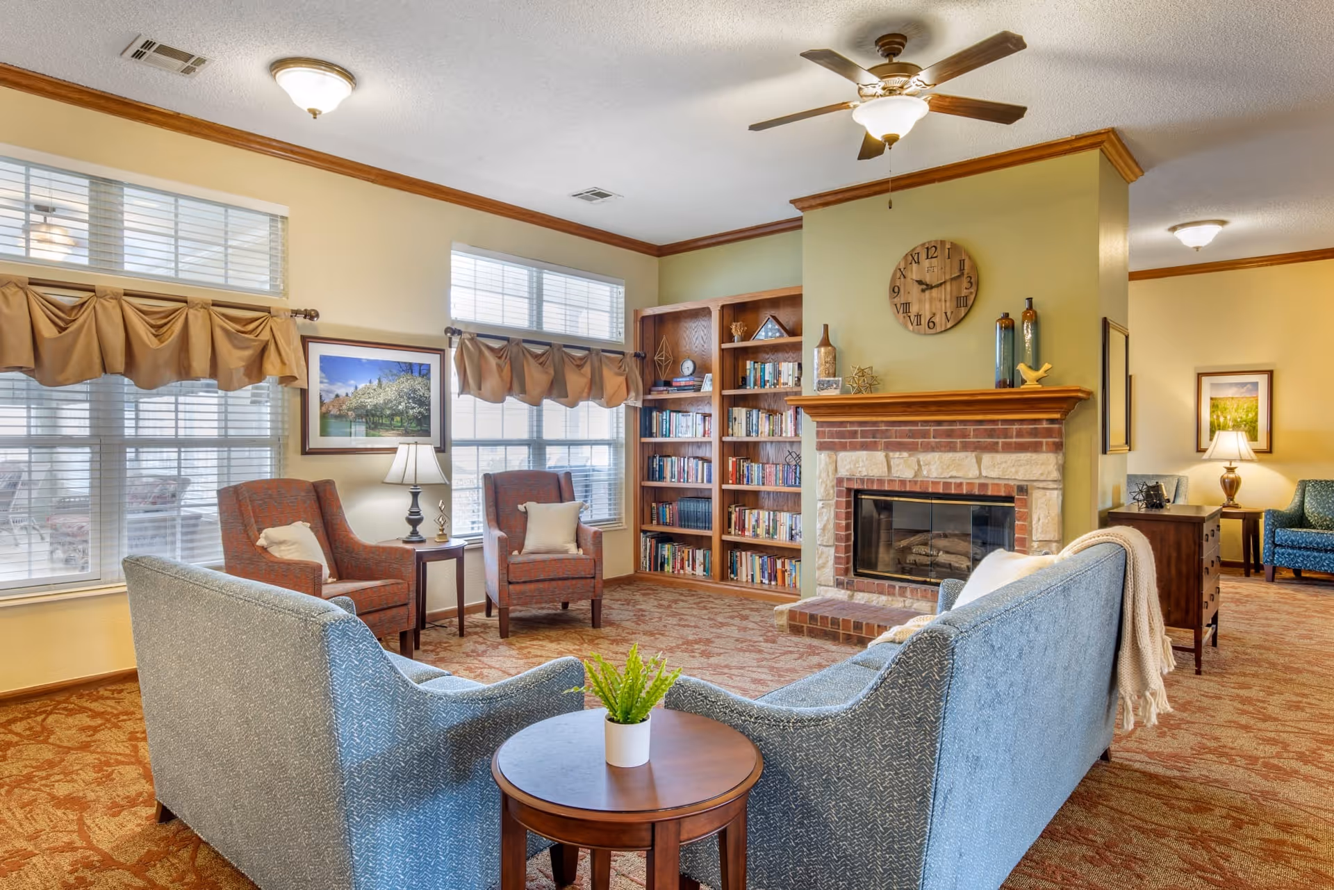 A cozy living room area with two blue upholstered sofas facing each other around a small round wooden table with a potted plant. Behind the sofas are two red armchairs with pillows and a side table with a lamp. The room features a brick and stone fireplace with decorative items on the mantel and a wooden clock above it. A built-in wooden bookshelf filled with books is next to the fireplace. The room has large windows with beige curtains and valances, and the floor is covered with a patterned carpet.