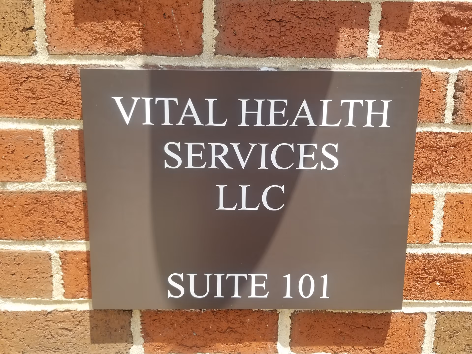 Brown metal sign reading Vital Health Services LLC, Suite 101, mounted on brick wall