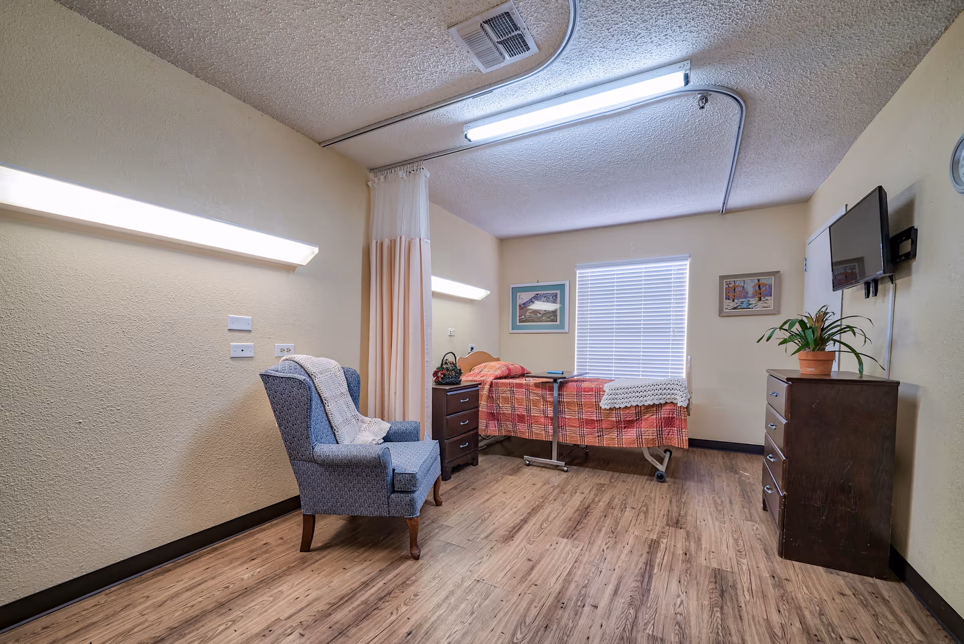 A nursing facility room with a single bed covered in a red plaid blanket, a blue upholstered armchair with a white throw draped over it, a wooden dresser with a potted plant, a wall-mounted TV, two framed pictures on the walls, and a window with blinds letting in natural light. The room has wood-look flooring and beige walls with fluorescent ceiling lights.