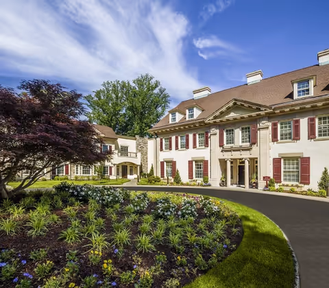 Exterior view of a large, elegant senior living facility building with beige walls, red shutters, and a brown roof. The building is surrounded by a well-maintained garden with various plants and flowers, and a curved driveway leading to the entrance. The sky is blue with some clouds.