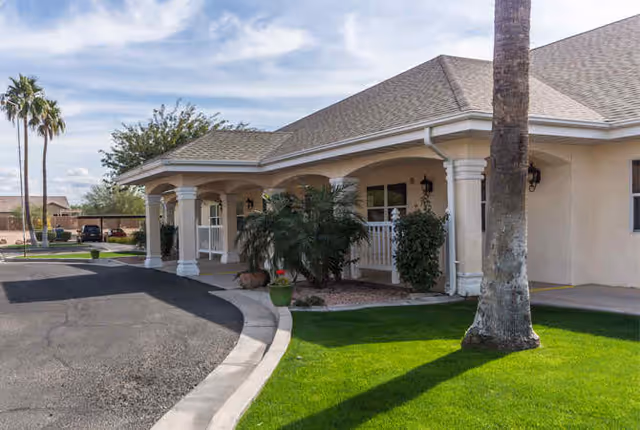 Front entrance of a single-story senior living building with a covered drive-up portico, columns, palm trees, and a green lawn.